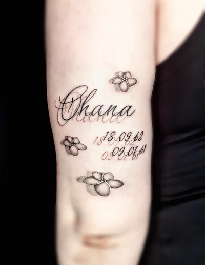 Schriftzug-Tattoo „Ohana“ mit drei feinen Blumen und zwei Daten in eleganter Schreibschrift auf dem Oberarm.