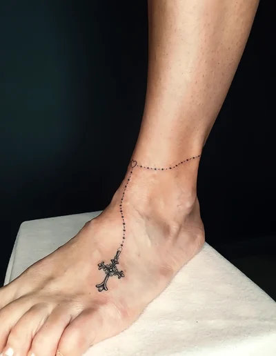 Realistic Tattoo eines filigranen Rosenkranzes mit Kreuz und Herz am Fußgelenk und Fuß.