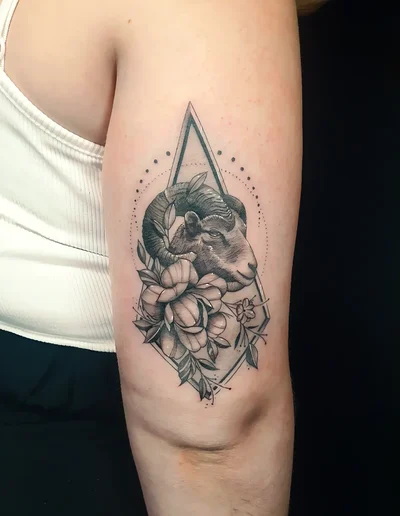 Realistic Tattoo eines Widderkopfs mit floralen Elementen und geometrischem Rahmen auf dem Oberarm.